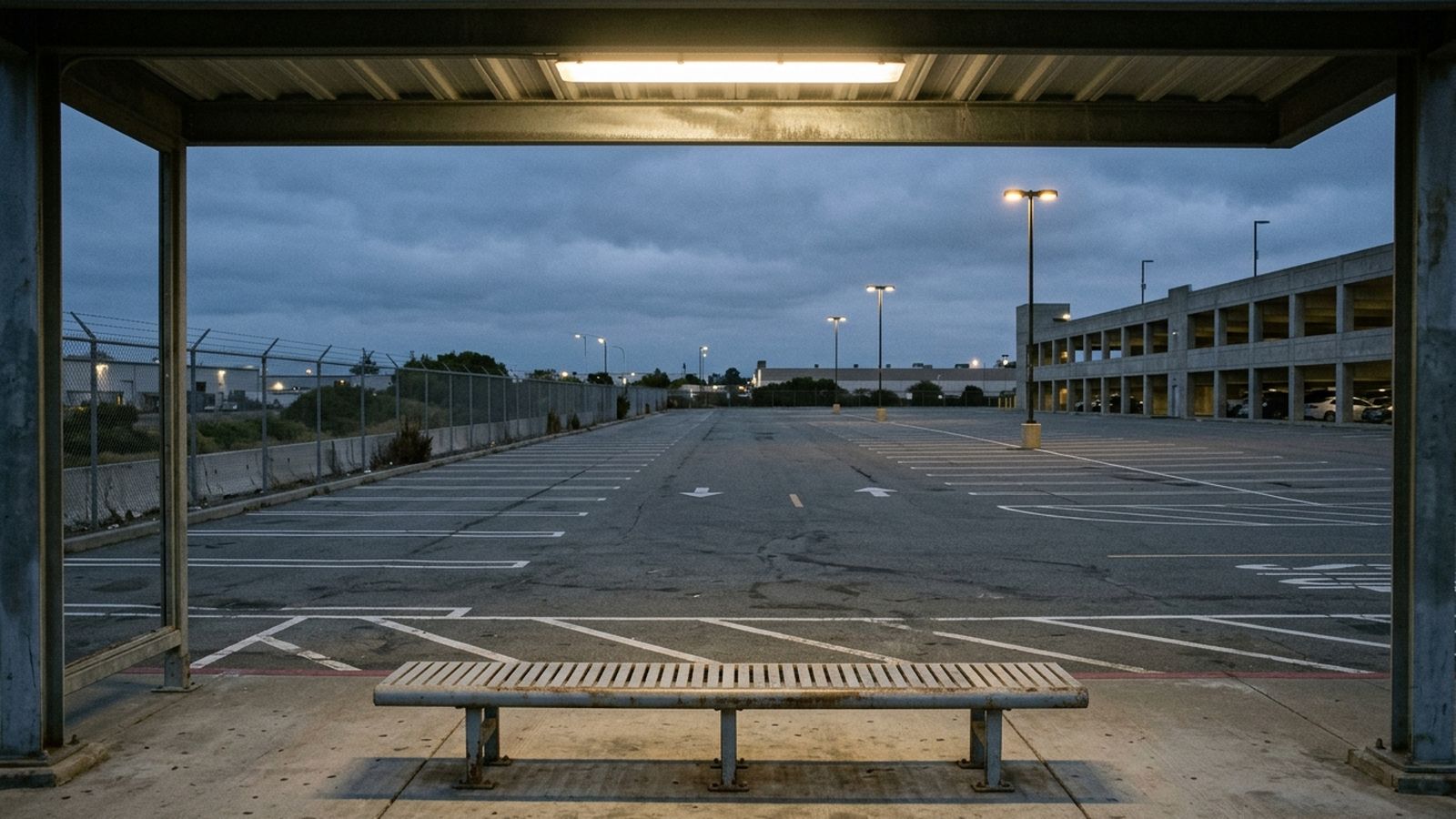 The inside of a covered shuttle shelter looking out over a large empty asphalt lot at dusk, a plain metal bench in the foreground and the overhead lights just coming on, nobody waiting